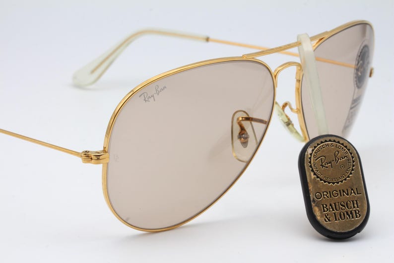 May include: Gold-tone aviator sunglasses with brown lenses and a Ray-Ban logo on the frame. The sunglasses are accompanied by a round, gold-tone metal tag with the text "Ray-Ban Original Bausch & Lomb".