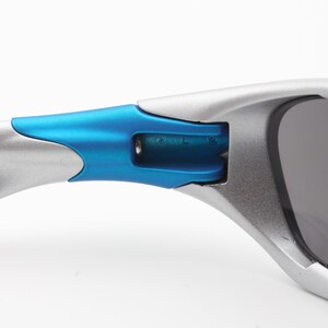May include: Close-up of a silver and blue pair of sunglasses. The sunglasses have a curved frame and dark lenses. The arms of the sunglasses are silver and blue.