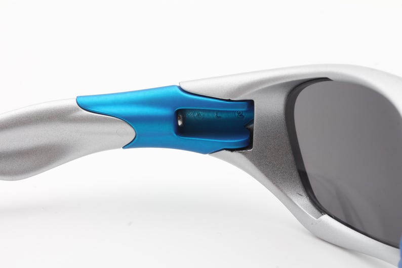 May include: Close-up of a silver and blue pair of sunglasses. The sunglasses have a curved frame and dark lenses. The arms of the sunglasses are silver and blue.