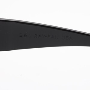 May include: Close-up of a black sunglasses arm with the text "B&L RAY-BAN USA" embossed on the surface. The sunglasses arm is curved and has a glossy finish. The background is white.
