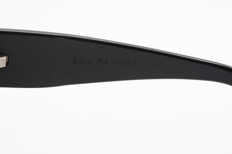 May include: Close-up of a black sunglasses arm with the text "B&L RAY-BAN USA" embossed on the surface. The sunglasses arm is curved and has a glossy finish. The background is white.