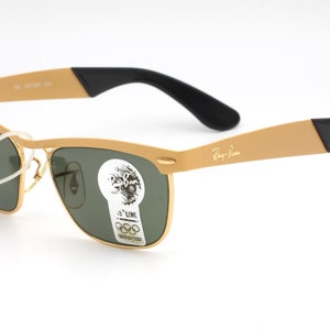 Puede incluir: Gafas de sol Ray-Ban doradas con lentes verdes y patillas negras. Las gafas tienen un estilo aviador cl&aacute;sico con un fino marco de metal.