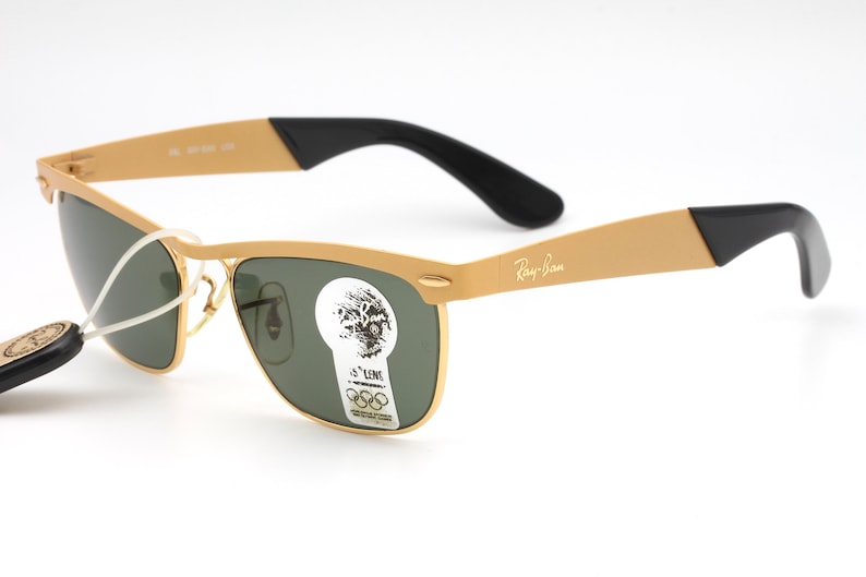 Puede incluir: Gafas de sol Ray-Ban doradas con lentes verdes y patillas negras. Las gafas tienen un estilo aviador cl&aacute;sico con un fino marco de metal.