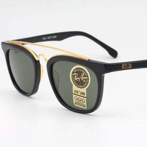 May include: Black Ray-Ban sunglasses with gold accents. The sunglasses have a classic design with a gold bridge and green lenses. The Ray-Ban logo is visible on the lens. The sunglasses are set against a white background.