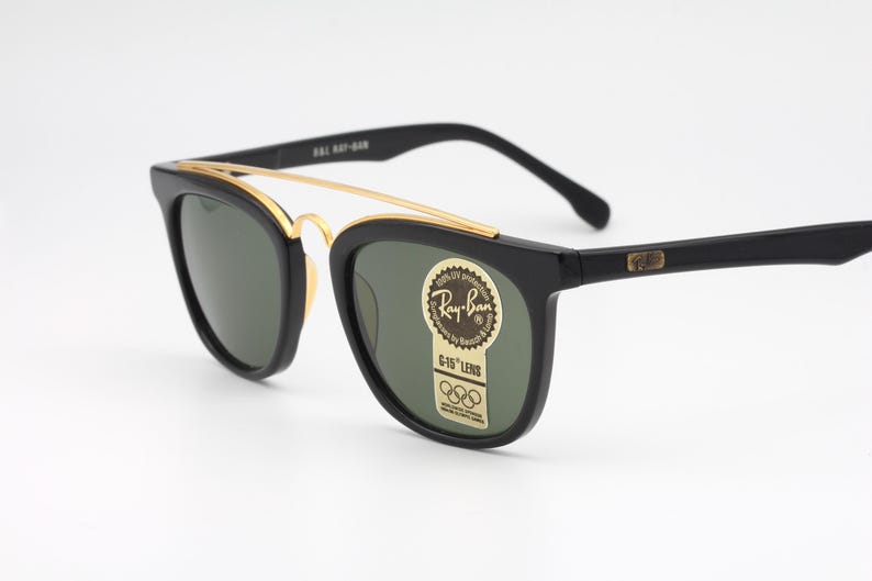 May include: Black Ray-Ban sunglasses with gold accents. The sunglasses have a classic design with a gold bridge and green lenses. The Ray-Ban logo is visible on the lens. The sunglasses are set against a white background.