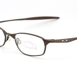 Puede incluir: Gafas de metal marrón con lentes transparentes. La montura tiene una curva sutil y un pequeño logotipo ovalado en el lateral.