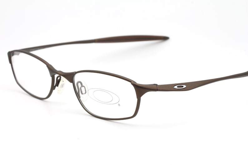May include: Brown metal frame spectacles with clear lenses. The frame has a subtle curve and a small oval logo on the side.