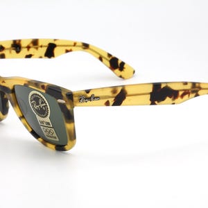 Puede incluir: Gafas de sol con estampado de carey y lentes oscuras. Las monturas y las patillas presentan un patr&oacute;n amarillo y marr&oacute;n. El logotipo de Ray-Ban es visible en la patilla derecha. Una pegatina con texto est&aacute; en la lente izquierda.