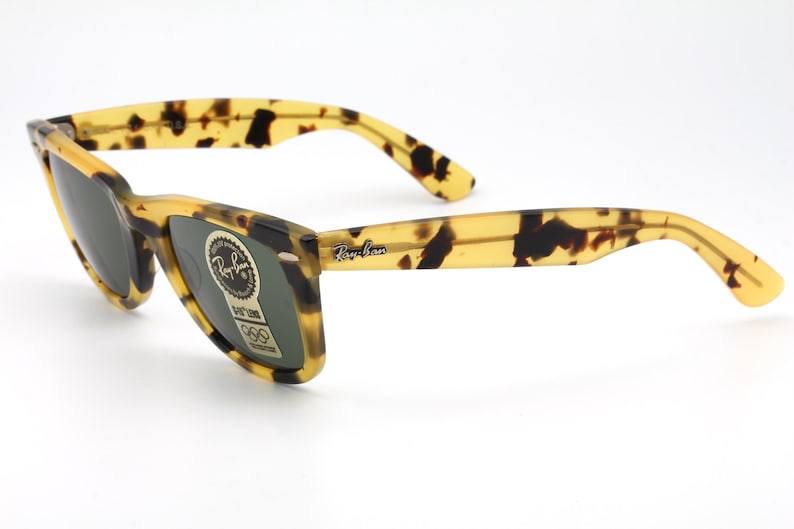 Puede incluir: Gafas de sol con estampado de carey y lentes oscuras. Las monturas y las patillas presentan un patr&oacute;n amarillo y marr&oacute;n. El logotipo de Ray-Ban es visible en la patilla derecha. Una pegatina con texto est&aacute; en la lente izquierda.