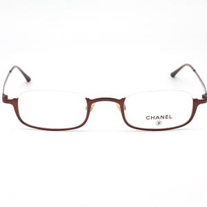 Puede incluir: Gafas con montura burdeos y lentes transparentes. Las gafas tienen un diseño de media montura con la marca "CHANEL" impresa en la lente derecha. Las patillas son de color marrón oscuro.