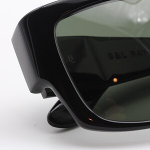 May include: Close-up of black sunglasses with dark green lenses. The sunglasses have a thick, rectangular frame and the text "B&L RAY" is visible on the lens. The sunglasses are set against a white background.