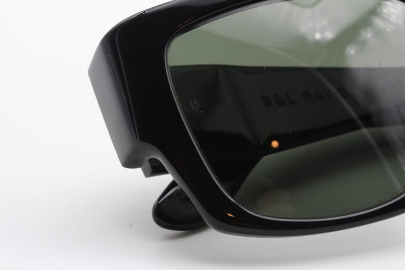 May include: Close-up of black sunglasses with dark green lenses. The sunglasses have a thick, rectangular frame and the text "B&L RAY" is visible on the lens. The sunglasses are set against a white background.