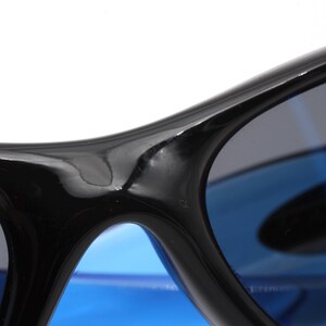 May include: Close-up of a pair of black sunglasses with blue lenses. The sunglasses have a curved frame and a glossy finish.