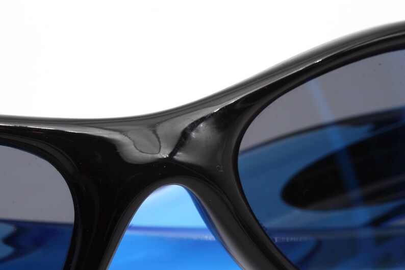 May include: Close-up of a pair of black sunglasses with blue lenses. The sunglasses have a curved frame and a glossy finish.
