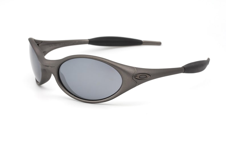 May include: Gray oval-shaped sunglasses with gray lenses. The frames are a matte gray color, with black rubber accents on the temples. The sunglasses have a sporty, streamlined design, suitable for outdoor activities.