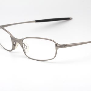 Puede incluir: Un par de gafas plateadas con montura rectangular y lentes transparentes. Las patillas tienen puntas negras. Las gafas están sobre un fondo blanco.