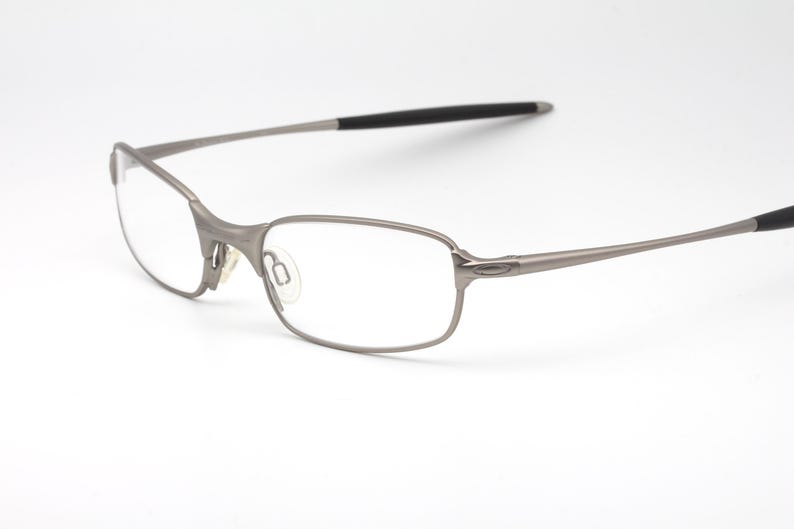 May include: A pair of silver-toned eyeglasses with a rectangular frame and clear lenses. The temples have black tips. The glasses are set against a white background.