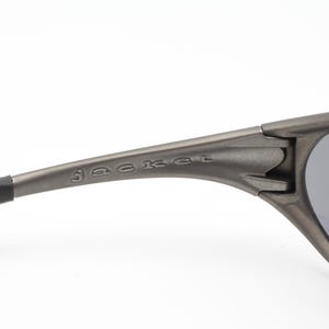 May include: Close-up of a pair of gray sunglasses with dark lenses. The word "jacket" is printed on the arm of the sunglasses. The sunglasses have a sleek, modern design.
