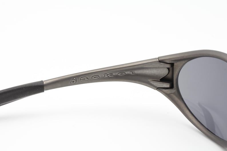 May include: Close-up of a pair of gray sunglasses with dark lenses. The word "jacket" is printed on the arm of the sunglasses. The sunglasses have a sleek, modern design.