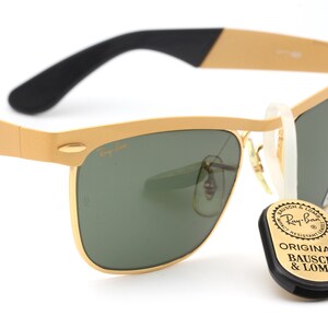 Puede incluir: Gafas de sol Ray-Ban doradas con lentes verdes y una varilla negra y dorada. Las gafas tienen un estilo aviador cl&aacute;sico con una montura de metal. Las gafas se acompa&ntilde;an de una etiqueta negra con el texto "Original Bausch & Lomb" en blanco.