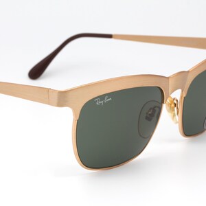 May include: Gold-tone Ray-Ban sunglasses with green lenses. The sunglasses have a square frame with a slight curve at the top.
