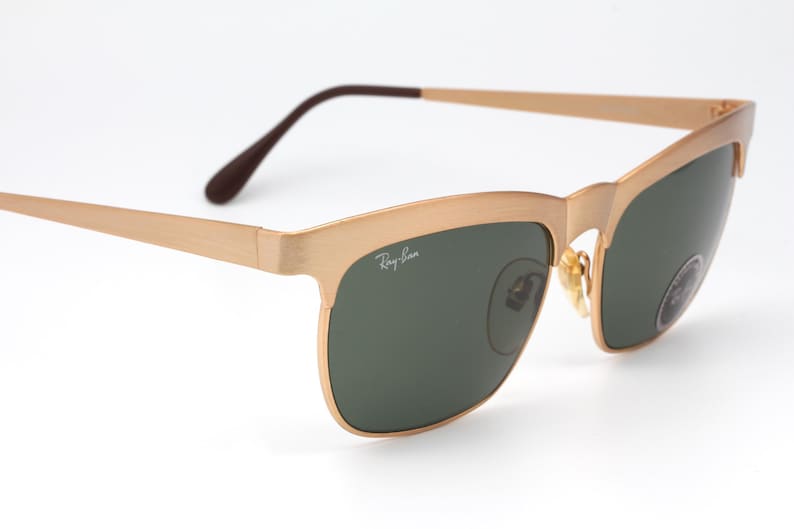May include: Gold-tone Ray-Ban sunglasses with green lenses. The sunglasses have a square frame with a slight curve at the top.