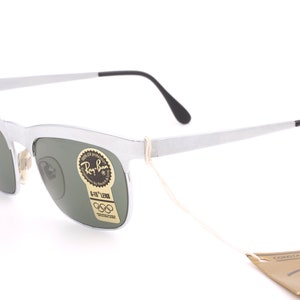 May include: Silver Ray-Ban sunglasses with green lenses and a G-15 lens tag. The sunglasses have a white string attached to them and a brown Ray-Ban tag with the text "CONTAIN GENUINE LENSES" and "Ray-Ban" in a white font.