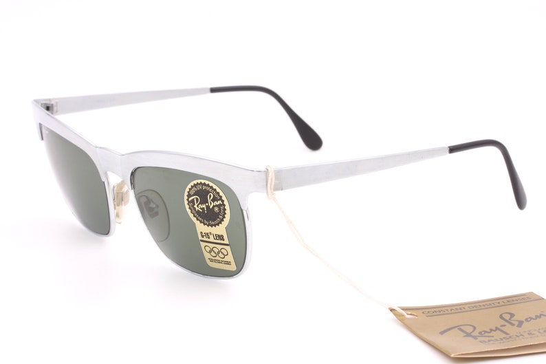 May include: Silver Ray-Ban sunglasses with green lenses and a G-15 lens tag. The sunglasses have a white string attached to them and a brown Ray-Ban tag with the text "CONTAIN GENUINE LENSES" and "Ray-Ban" in a white font.