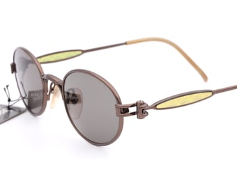 May include: Brown metal framed sunglasses with round lenses and a yellow accent on the arms. The sunglasses have a hinge with a unique design.