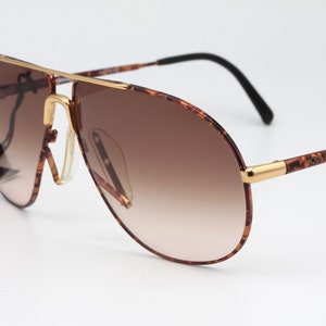 May include: Brown and gold aviator sunglasses with a tortoise shell frame and gradient brown lenses. The sunglasses have a gold metal bar across the bridge of the nose and the brand name "Vogue" is printed on the right arm.