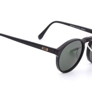 May include: Black round sunglasses with green lenses and a gold Ray-Ban logo on the side. The sunglasses have a black plastic frame and are attached to a white string.
