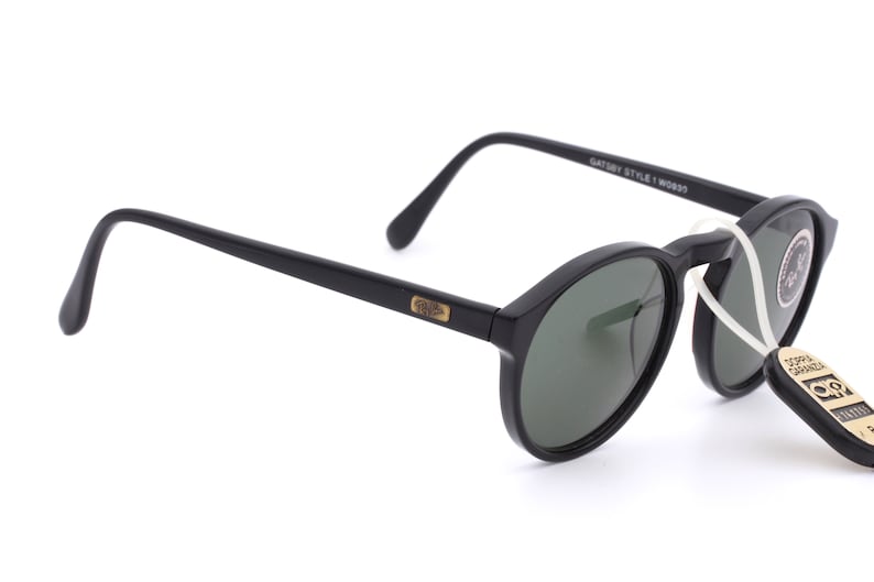 May include: Black round sunglasses with green lenses and a gold Ray-Ban logo on the side. The sunglasses have a black plastic frame and are attached to a white string.