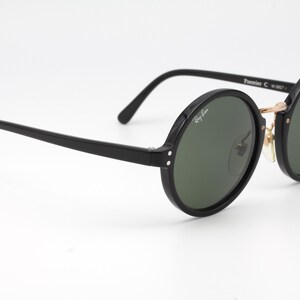 May include: Black round Ray-Ban sunglasses with green lenses. The sunglasses have a gold metal bridge and black plastic frames.