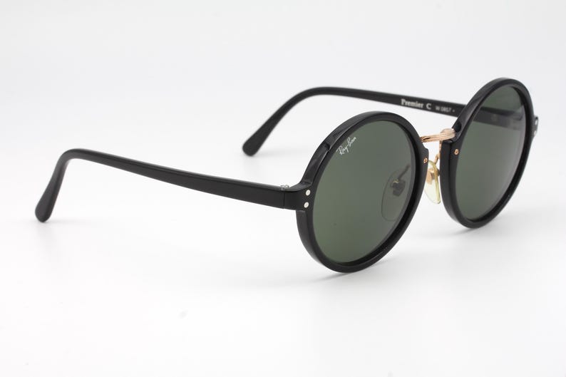 May include: Black round Ray-Ban sunglasses with green lenses. The sunglasses have a gold metal bridge and black plastic frames.