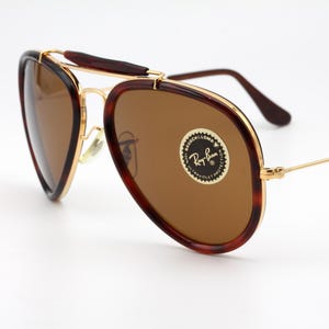 May include: Vintage Ray-Ban sunglasses with brown lenses and a gold frame. The frames have a tortoiseshell pattern on the outer edges. The lenses have a circular sticker with the Ray-Ban logo.