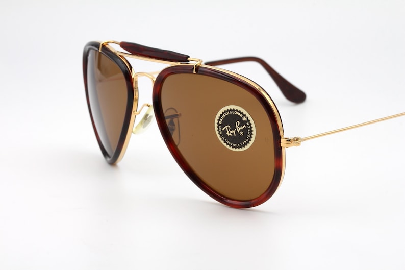 May include: Vintage Ray-Ban sunglasses with brown lenses and a gold frame. The frames have a tortoiseshell pattern on the outer edges. The lenses have a circular sticker with the Ray-Ban logo.