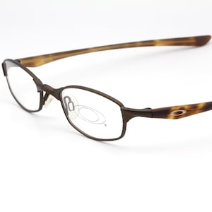 May include: Brown oval-framed eyeglasses with a tortoise shell pattern on the temples. The glasses have a sleek, modern design and a small logo on the temple. The lenses are clear, and the frame is a dark brown color.