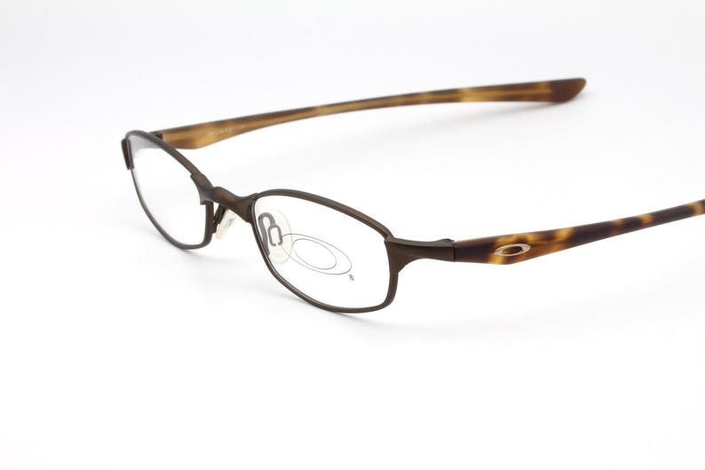 May include: Brown oval-framed eyeglasses with a tortoise shell pattern on the temples. The glasses have a sleek, modern design and a small logo on the temple. The lenses are clear, and the frame is a dark brown color.