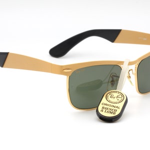 Puede incluir: Gafas de sol Ray-Ban doradas con lentes verdes y detalles negros. Las gafas tienen un estilo aviador cl&aacute;sico con una montura de metal y un puente de nariz de pl&aacute;stico negro. Las gafas vienen acompa&ntilde;adas de una peque&ntilde;a etiqueta negra que dice "Original Bausch & Lomb".