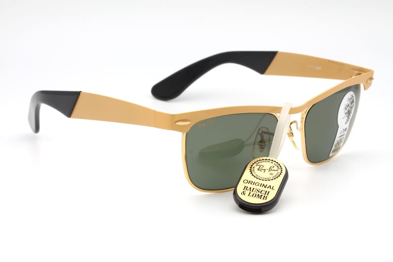 Puede incluir: Gafas de sol Ray-Ban doradas con lentes verdes y detalles negros. Las gafas tienen un estilo aviador cl&aacute;sico con una montura de metal y un puente de nariz de pl&aacute;stico negro. Las gafas vienen acompa&ntilde;adas de una peque&ntilde;a etiqueta negra que dice "Original Bausch & Lomb".
