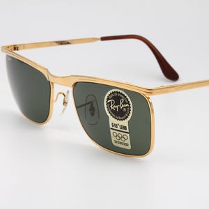 Op de afbeelding: Goudkleurige Ray-Ban zonnebril met donkergroene glazen. De bril heeft een rechthoekige vorm en bruine uiteinden van de pootjes. Een sticker op de lens leest "Ray-Ban" en "G-15 Lens" met Olympische ringen.