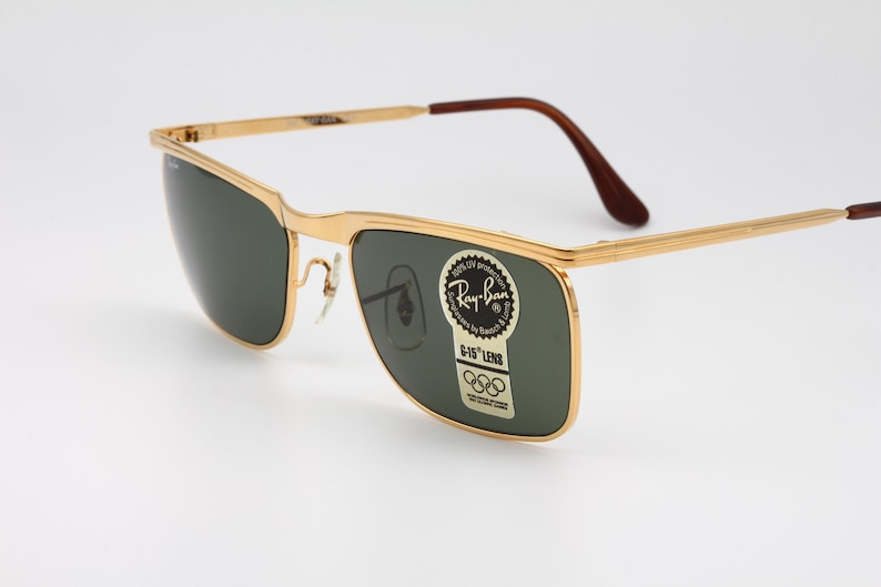 Op de afbeelding: Goudkleurige Ray-Ban zonnebril met donkergroene glazen. De bril heeft een rechthoekige vorm en bruine uiteinden van de pootjes. Een sticker op de lens leest "Ray-Ban" en "G-15 Lens" met Olympische ringen.