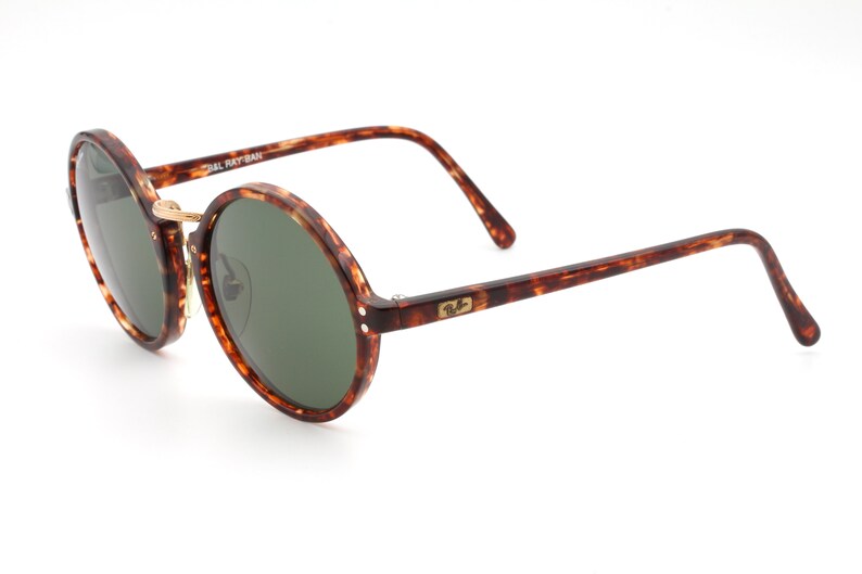 May include: Tortoiseshell round frame sunglasses with green lenses. The sunglasses have a gold metal bridge and the Ray-Ban logo is visible on the right arm.