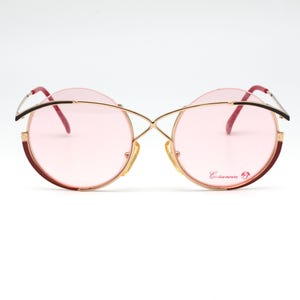 May include: Vintage round sunglasses with pink lenses and a gold-tone metal frame. The frames have a unique cross-over design and burgundy accents. The lenses have a subtle pink hue, and the arms are black and gold.