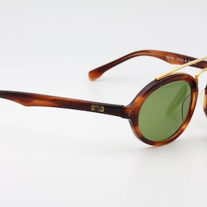 May include: Tortoise shell Ray-Ban sunglasses with green lenses and a gold metal frame. The sunglasses have a round shape and a classic design. The Ray-Ban logo is visible on the side of the frame. The sunglasses are accompanied by a small black tag that reads "Original Bausch & Lomb".