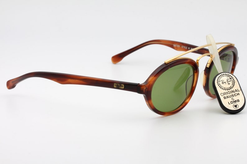 May include: Tortoise shell Ray-Ban sunglasses with green lenses and a gold metal frame. The sunglasses have a round shape and a classic design. The Ray-Ban logo is visible on the side of the frame. The sunglasses are accompanied by a small black tag that reads "Original Bausch & Lomb".