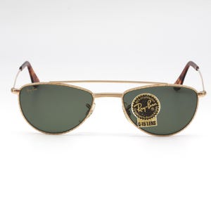 May include: Gold-framed Ray-Ban sunglasses with green lenses. The sunglasses have a classic aviator style with a double bridge and brown tortoiseshell temples. A Ray-Ban sticker is on the lens.