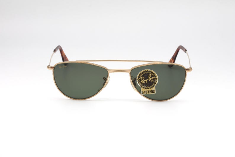 May include: Gold-framed Ray-Ban sunglasses with green lenses. The sunglasses have a classic aviator style with a double bridge and brown tortoiseshell temples. A Ray-Ban sticker is on the lens.