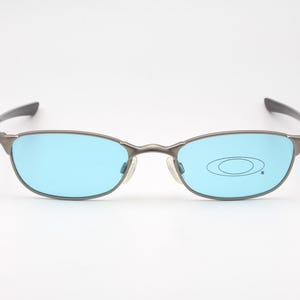 May include: Gray metal-framed sunglasses with light blue lenses. The oval-shaped lenses are set in a thin, rectangular frame with a subtle nose bridge and dark gray temples. The Oakley logo is visible on the right lens.