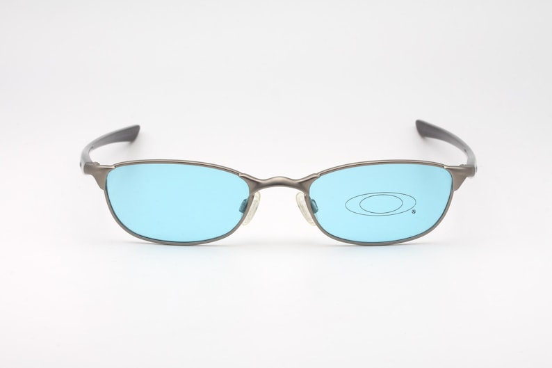 May include: Gray metal-framed sunglasses with light blue lenses. The oval-shaped lenses are set in a thin, rectangular frame with a subtle nose bridge and dark gray temples. The Oakley logo is visible on the right lens.